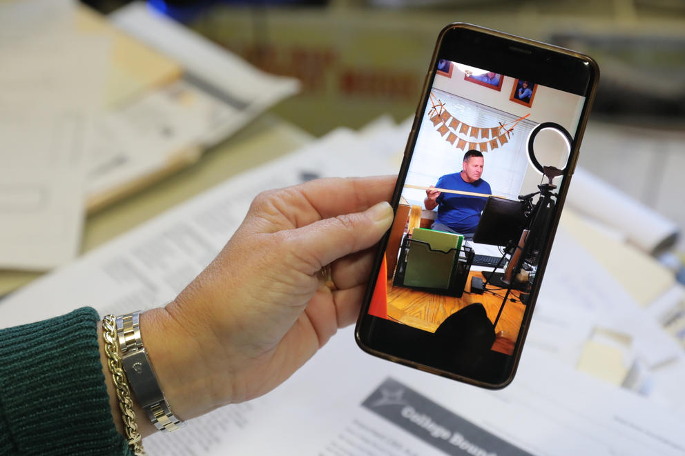 A hand holds a phone with a video of a man standing behind a ring light and a computer holding a piece of wood