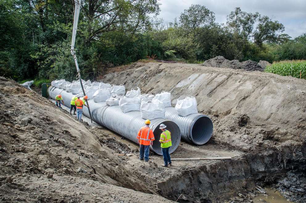 Workers in an excavated area stand by two large tubes, which will serve as fish passageways, that are held down with sandbags