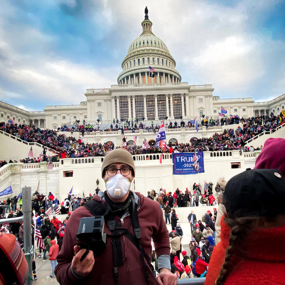 Person in brown hat and maroon sweater, wearing glasses and an N95  mask in front of the Capitol, in a color photo