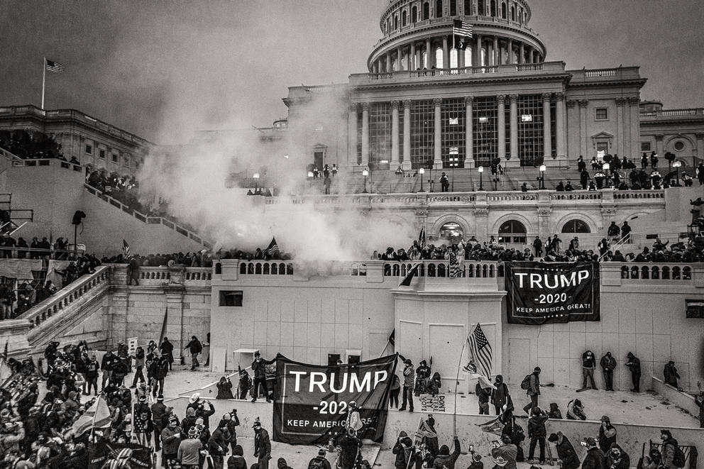 The capitol building is seen from afar, with the steps and platforms visible, there are people and flags and there is smoke from teargass, in a black and white photo