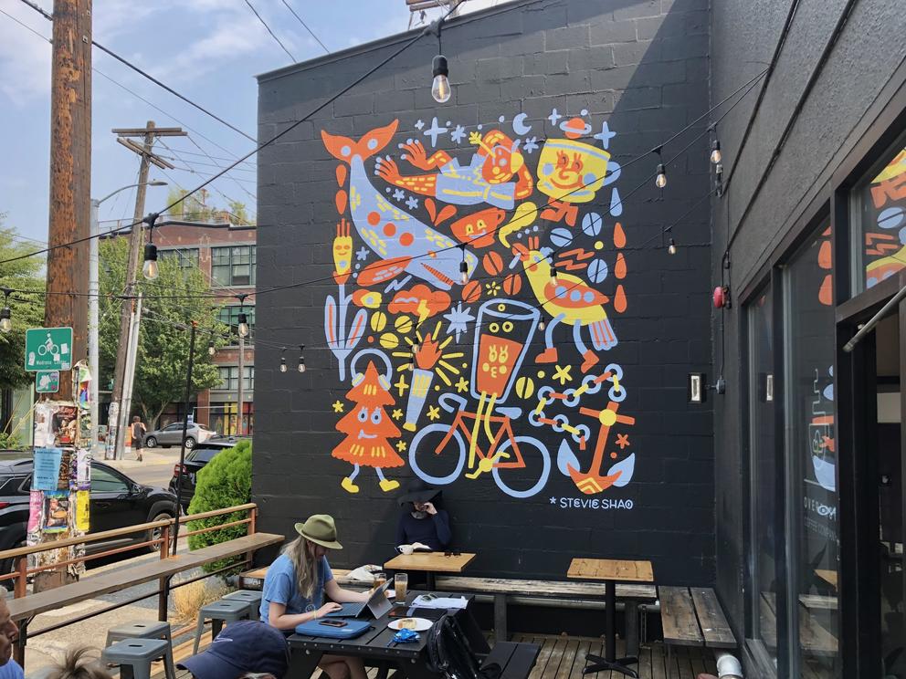 Cafe with person sitting behind laptop, outside, in front of a tall black wall featuring a colorful mural with animals and bikes.