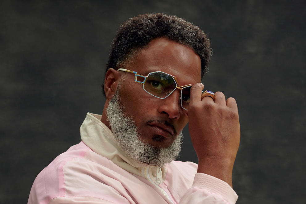 A bearded man looks pensively at the camera as he wears geometric sunglasses