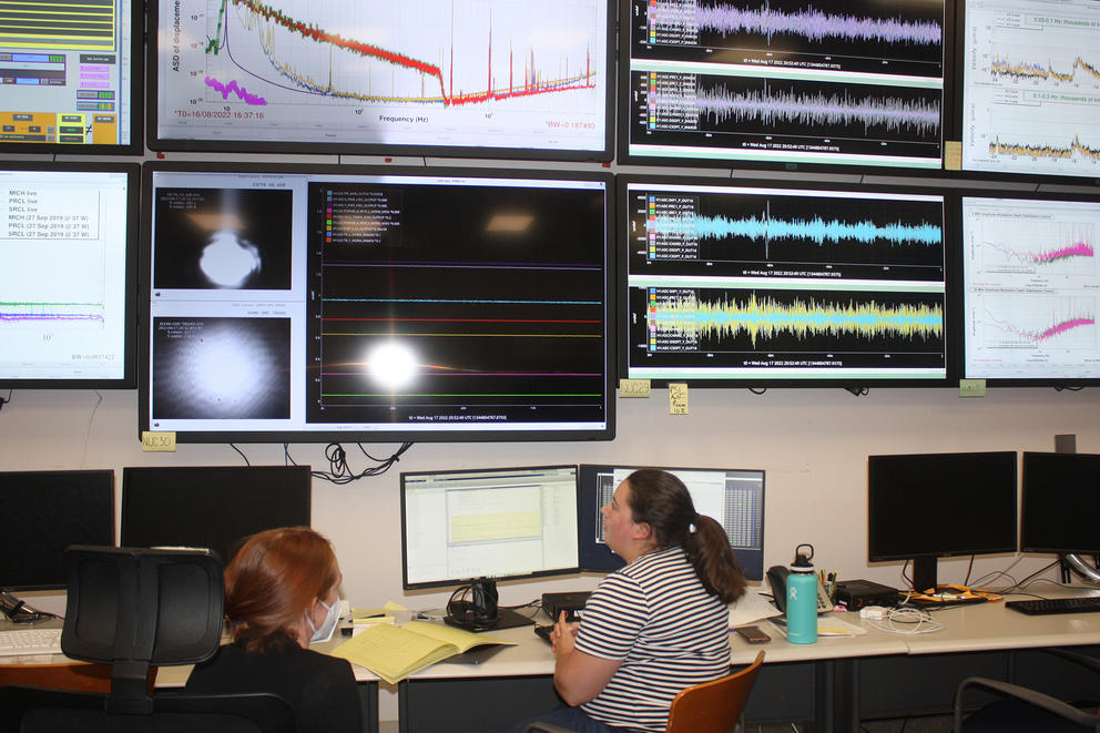 Two people sitting at desks facing a wall of large screens depicting wavelengths and charts