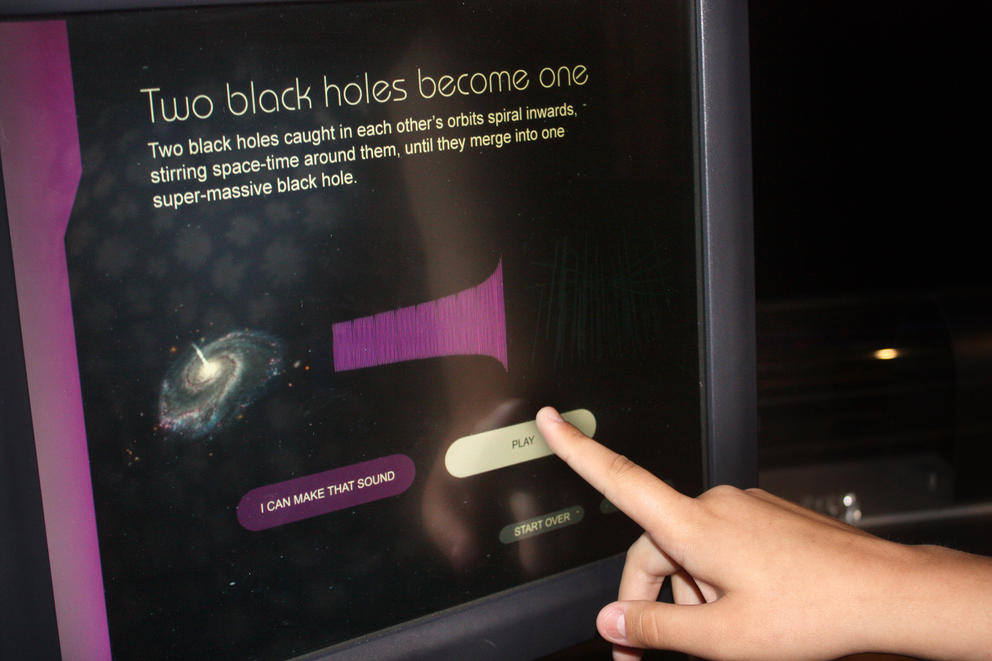 A computer screen depicts a black hole. The screen reads: "Two black holes become one — Two black holes caught in each other's orbits spiral inwards, stirring space-time around them, until they merge into one super-massive black hole." Buttons read: "I can make that sound" and "Play"