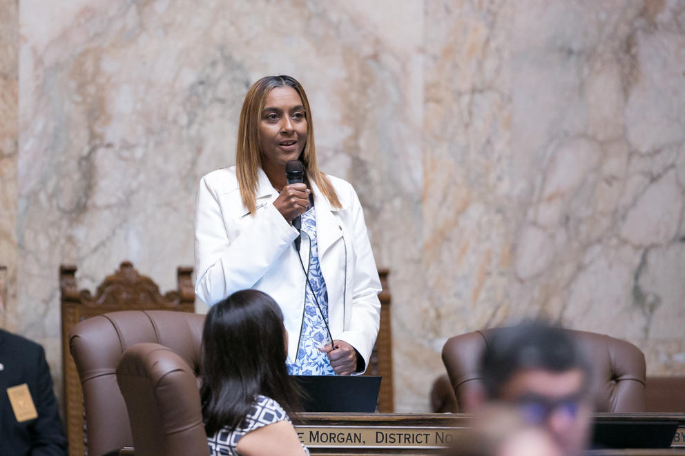 State Rep. Melanie Morgan holds a microphone as she speaks on the floor of the state House, marble walls behind her