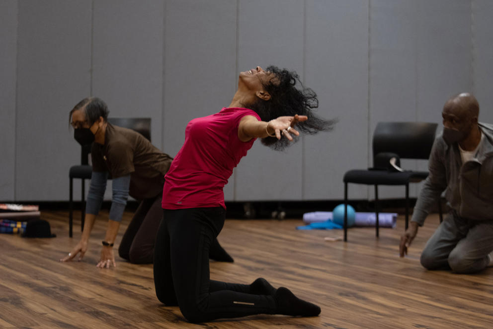 A woman kneeling on the floor with her head and arms thrown back, hair flying, during a dance class