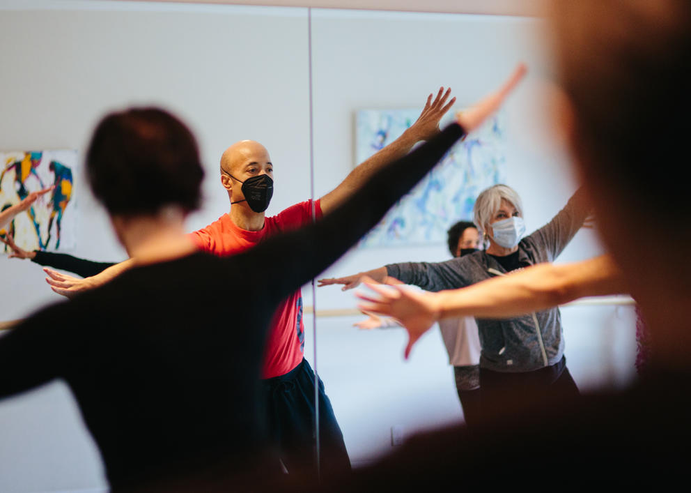 Chris Daigre is seen wearing a mask and red shirt through the raised arms of students in his dance class