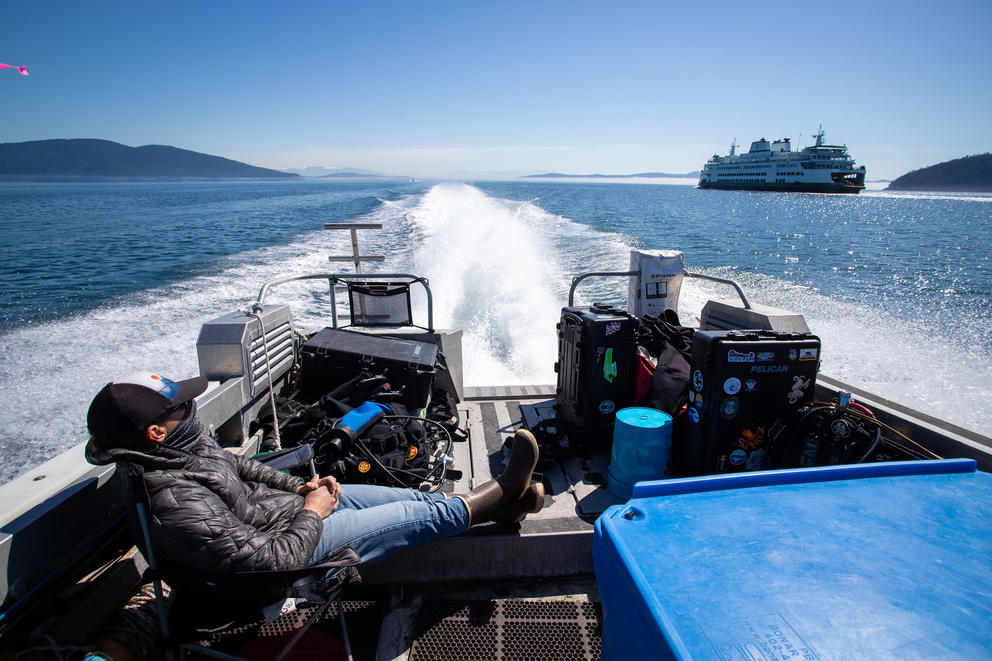 A man sits with his feet up at the back of a moving boat, a ferry passes by in the background