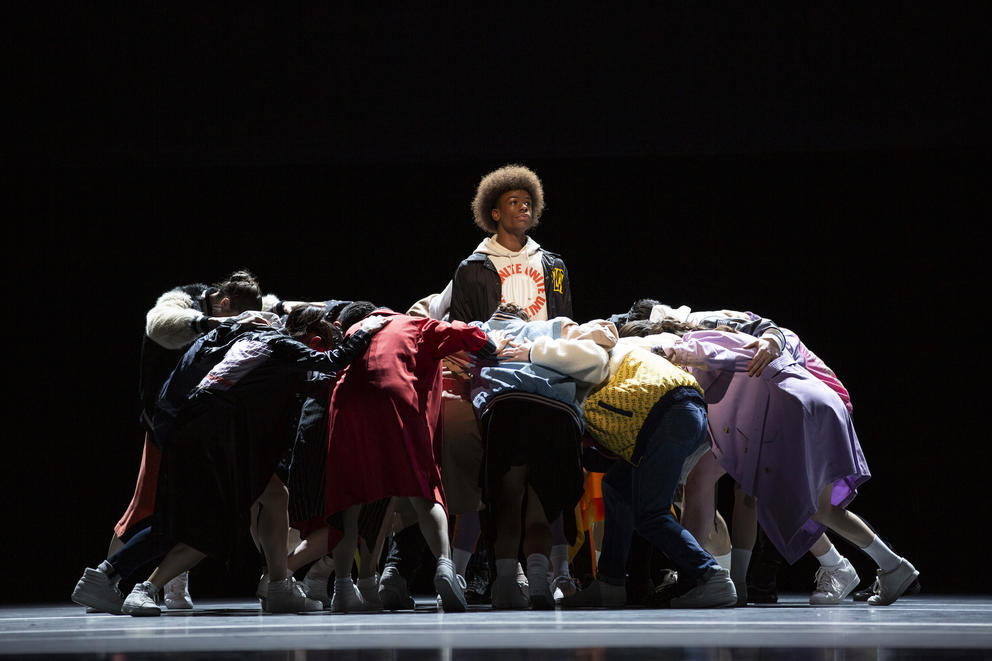 a photo of dancers performing on stage, one young man at the center of many others 