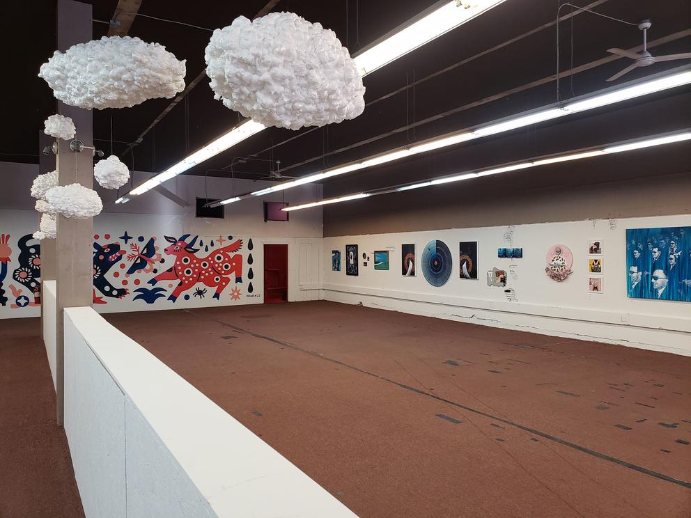 A large space with brown floors with overhead bright lights. On the wall left is a large mural of mythical animals in a neo-naive style, on the right hang multiple paintings and artworks