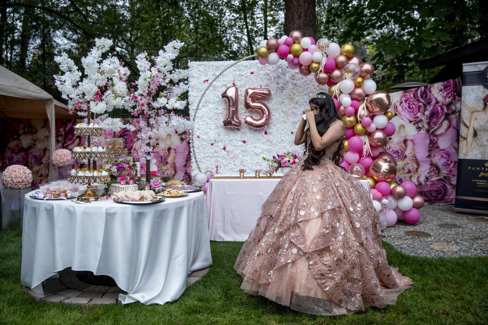 Evelyn Hernandez is wearing a pink gown and surrounded by pink and gold balloons and decorations as she wipes away a tear at her quinceañera 