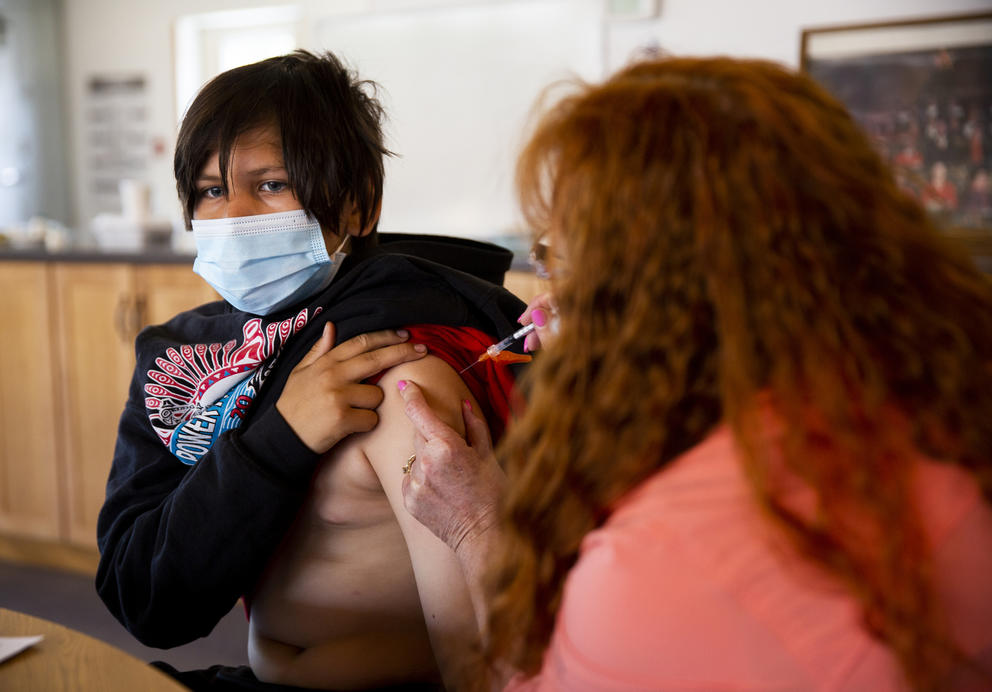 Barbara Hoffman administers a vaccine to Urijah Woodward who is wearing a mask and  has his sweatshirt pulled up over his shoulder 