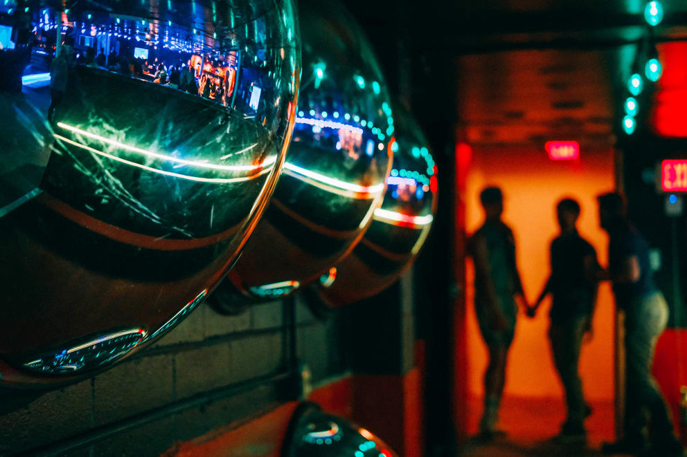 Two shadowy figures hold hands in the background, in the foreground convex mirrors reflect a the interior of Lumber Yard bar