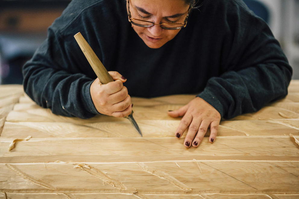 Andrea Wilbur-Sigo is bent over a large cedar log with a chisel in her hand carving designs into the wood