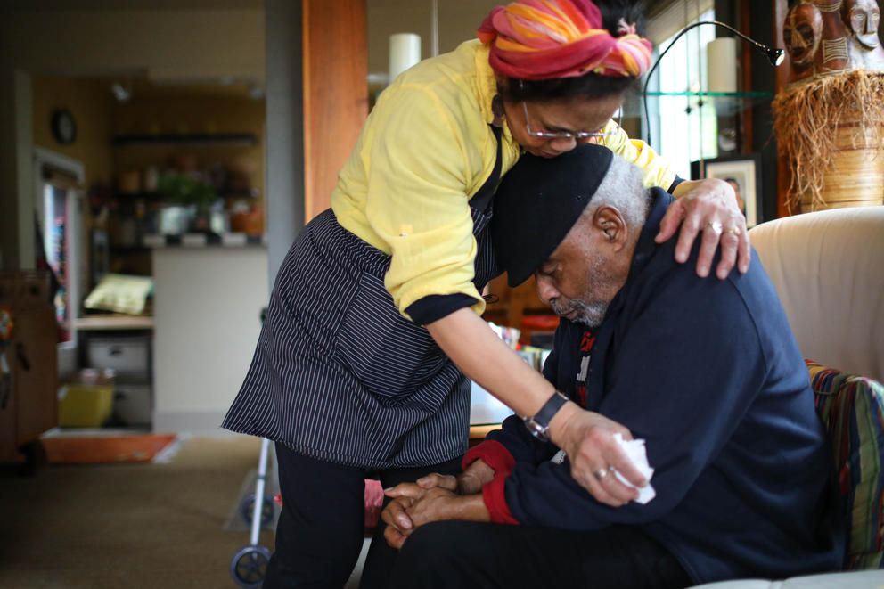 A woman stands over a man and wraps her arms around him, her head rests on his