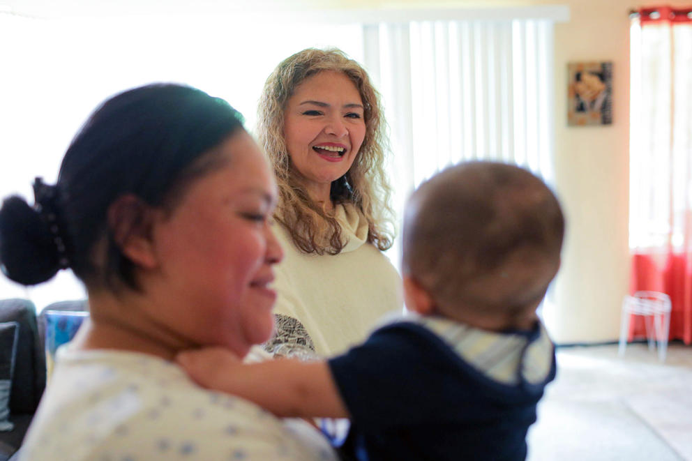 A woman holds a baby, a second woman is seen between them facing the camera and smiling at the baby