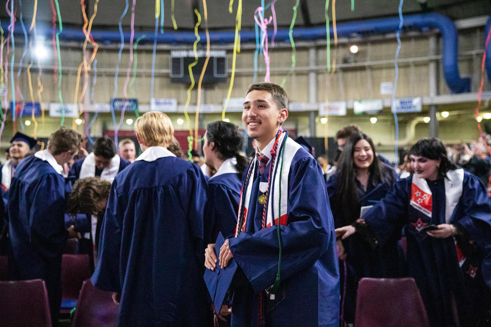 Jordan Chaves smiles in a crowd of his fellow graduates, wearing blue robes. Confetti falls from above.