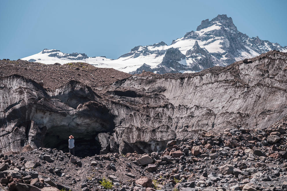 A snowy mountain peak rises in the background above a rock field. A small figure is dwarfed on the rock field in the distance.