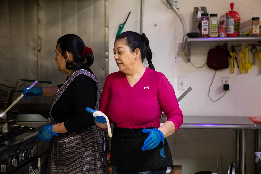 Two woman cook shoulder-to-shoulder in a stainless steel commercial kitchen.