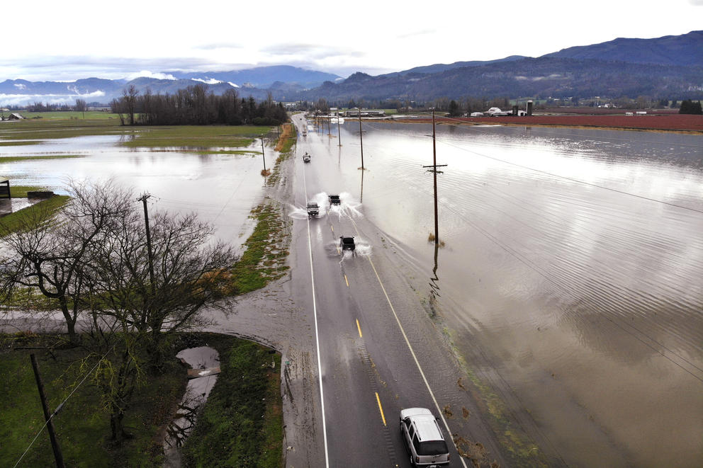 Trucks drive through shallow flood water encroaching on a two-lane highway.