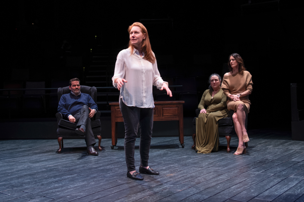 Person in a white shirt stands up in a dark room, three people sit behind her