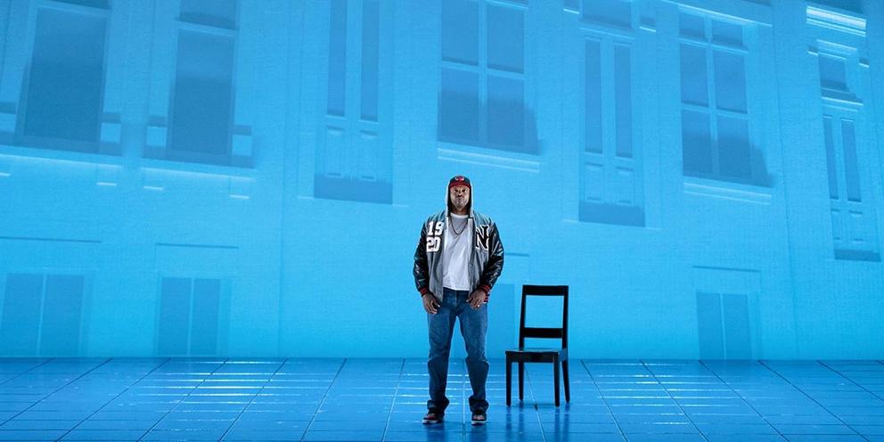 a stage scene featuring a black man in baseball cap standing in front of a blue wall of buildings