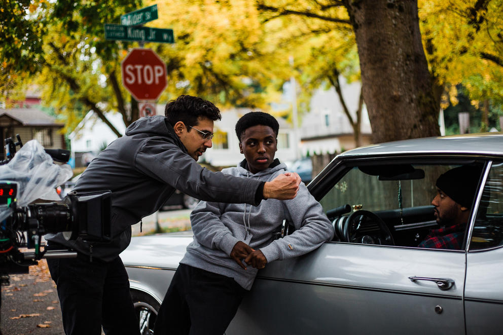 film still with a director in a black jacket directing a young actor leaning against a car