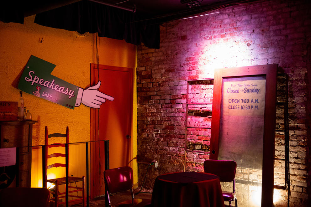 A two-seat table in the corner of the theatre. A sign in the shape of an arm pointing to the right reads "Speakeasy - shhh..."