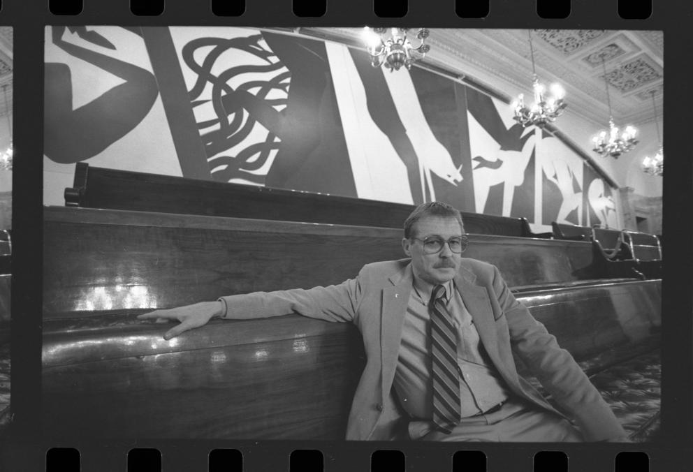 black and white photo of a man in a suit sitting on a bench in front of a large mural