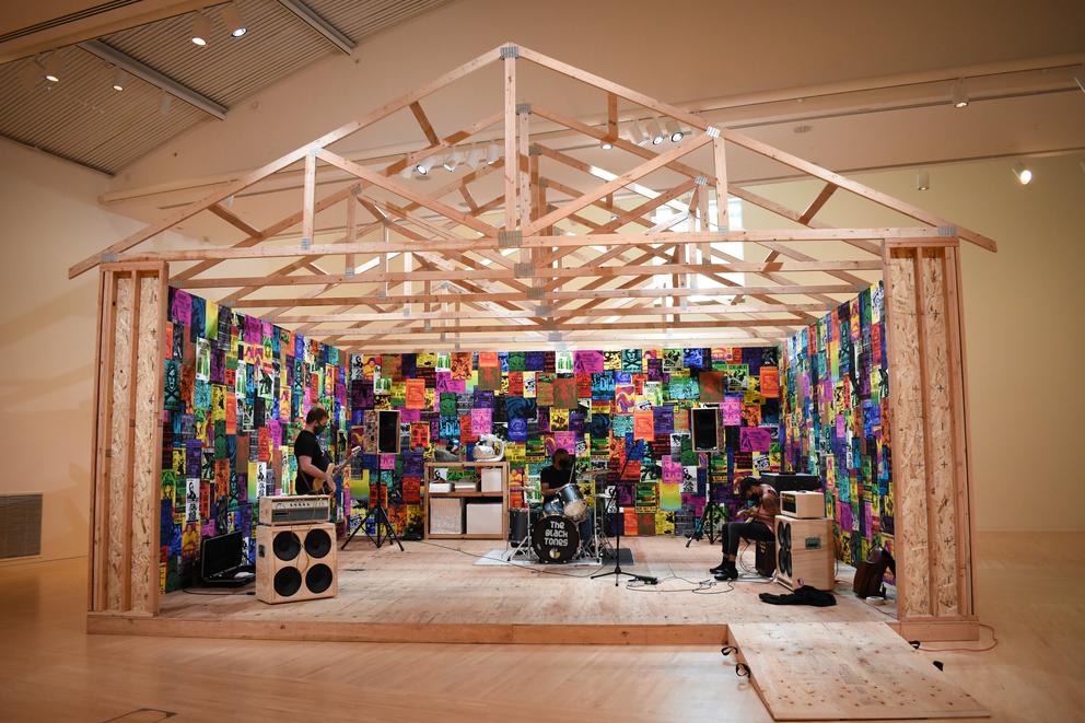 Plywood shed with wheatpasted posters inside, the frame structure of the roof are just exposed beams. Inside, three musicians are seen rehearsing, guitars in hand, and one behind drums in the middle.