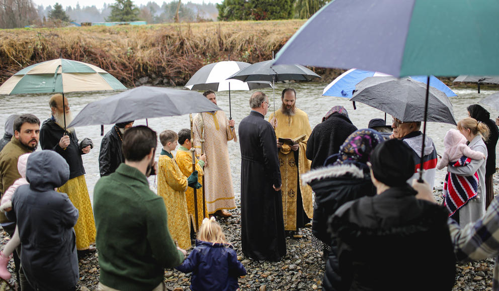 A group of people gather under umbrellas on a river bank around a priest in gold vestments