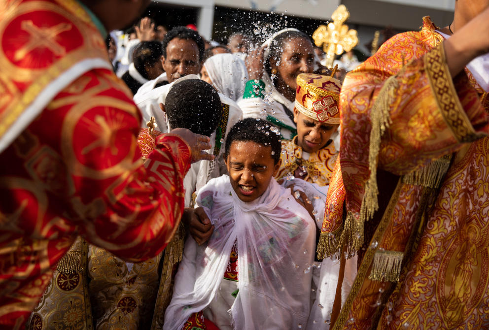 Priests sprinkle water on congregants in white robes, a young boy squints as water hits his face