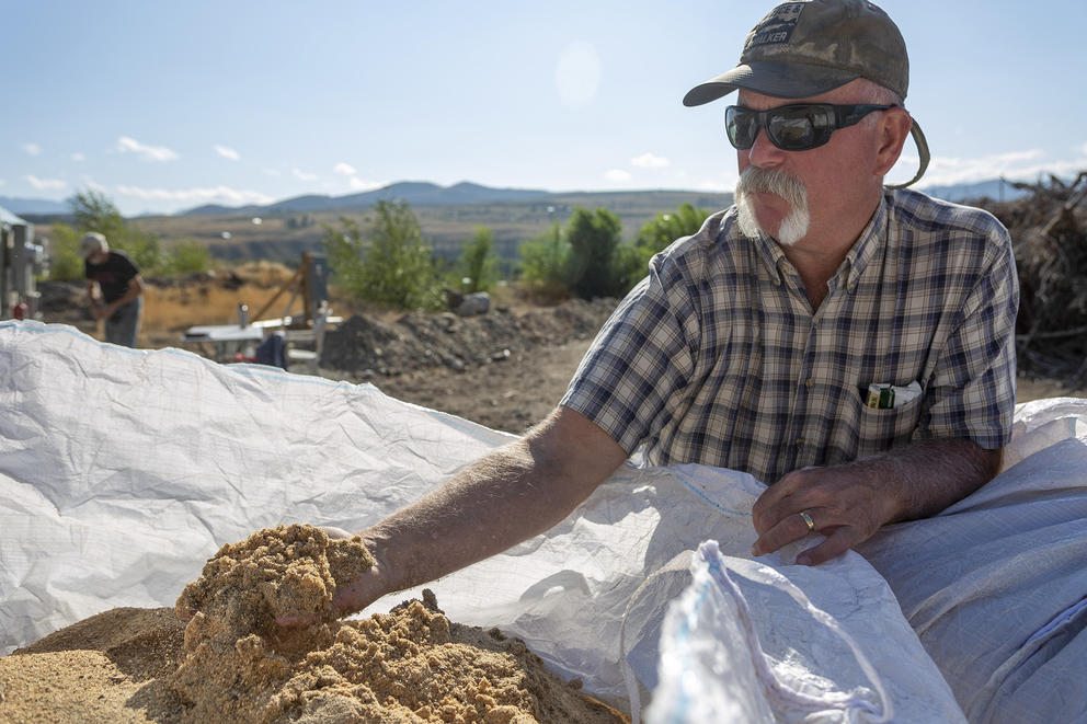 A man in a hat and sunglasses cups gold-toned dirt in his right hand