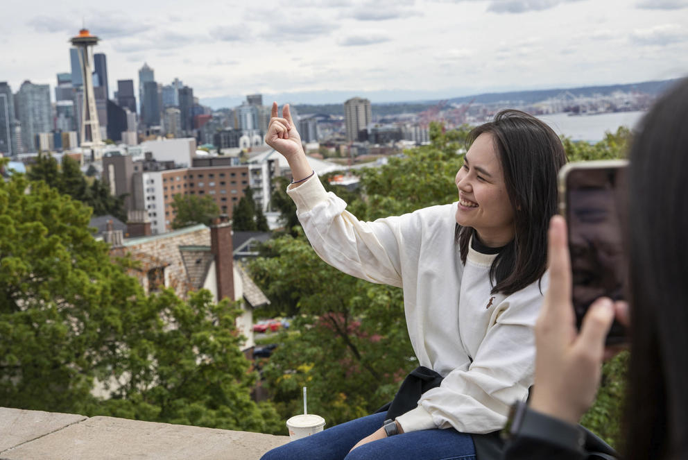 A person takes a photo of a woman with the Seattle skyline in the background