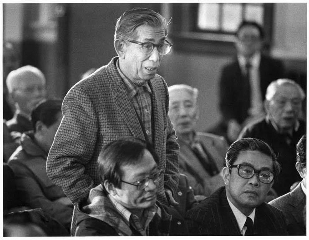 A man wearing glasses stands up and speaks, surrounded by other seated men