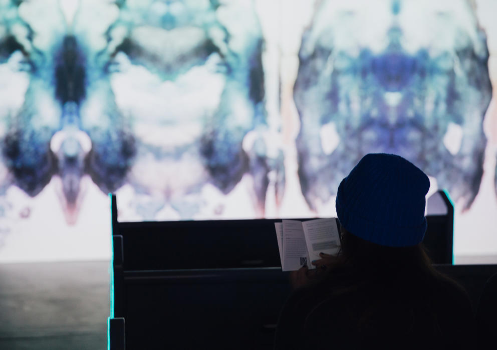 Person in right hand lower corner reading a book sitting on a pew while a screen fills the rest of the frame, Rorschach-like images in shades of blue are visible on the screen.