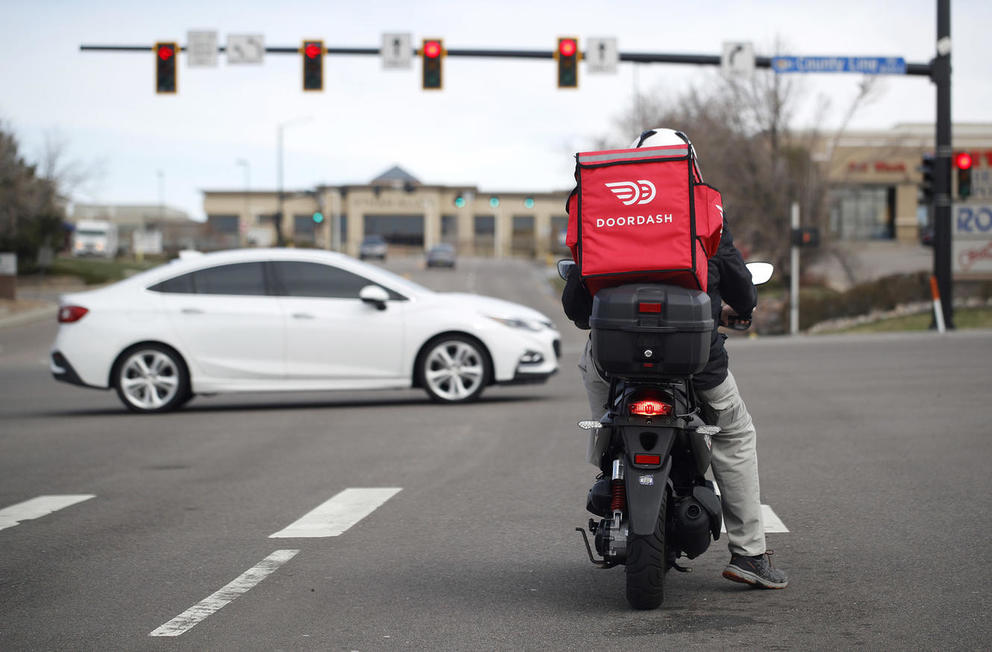 a doordash delivery person sits on a moped with a red food delivery bag on their back at a stop light on the street