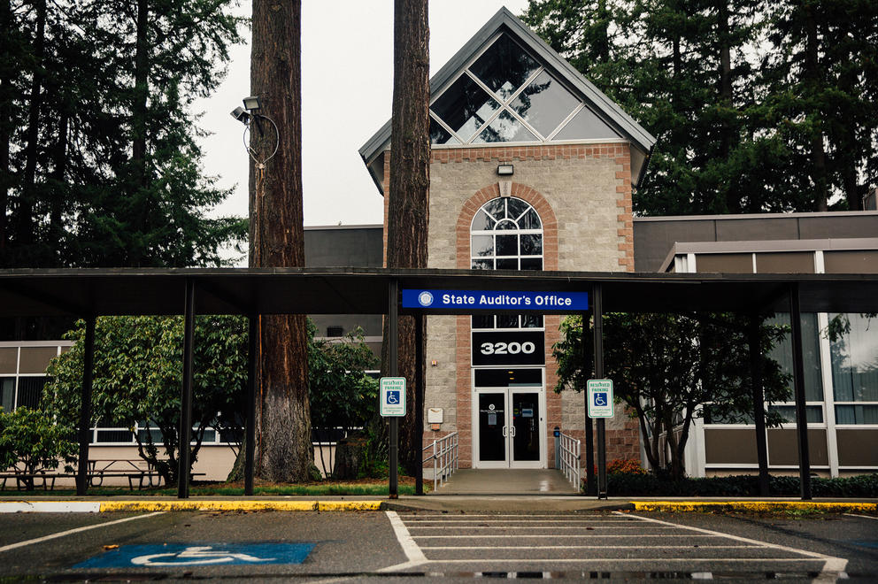 A blue sign reading State Auditor's Office hangs in front of an office building. 