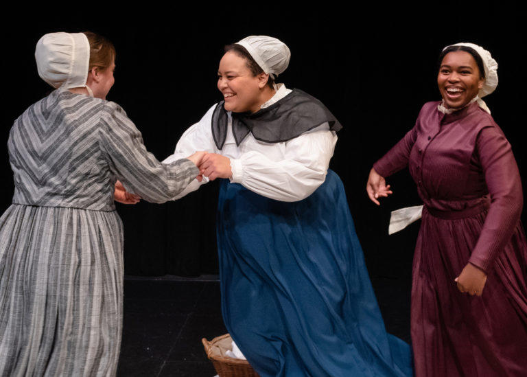 Two women dressed in Shaker dresses and bonnets hold each others hands while a third smiles