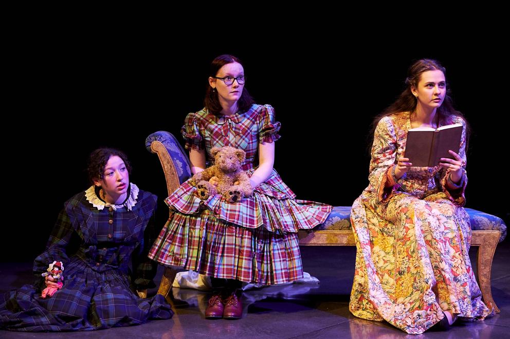 Three girls sit next to each other, one on the floor and two on a couch. They're dressed in costumes. One is holding an open book