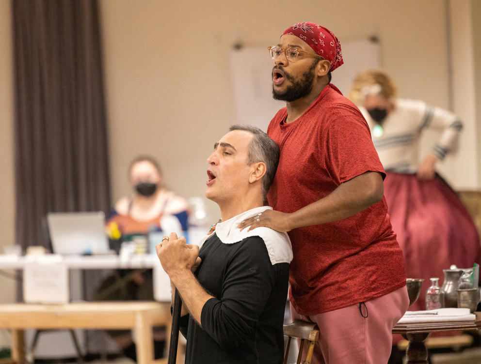 photo of a backstage rehearsal a man sits in front of another standing man, both singing