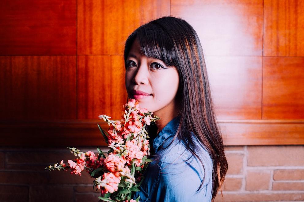 a woman with long brown hair turns back to look at the camera, with flowers in her hand