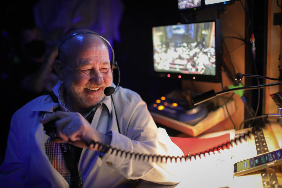 Production stage manager Jack McLeod smiles as he radios announcements to cast and crew. Behind him video feeds of the stage and audience are visible 