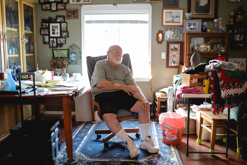 a man sits on a desk chair in front of a window in a cluttered apartment