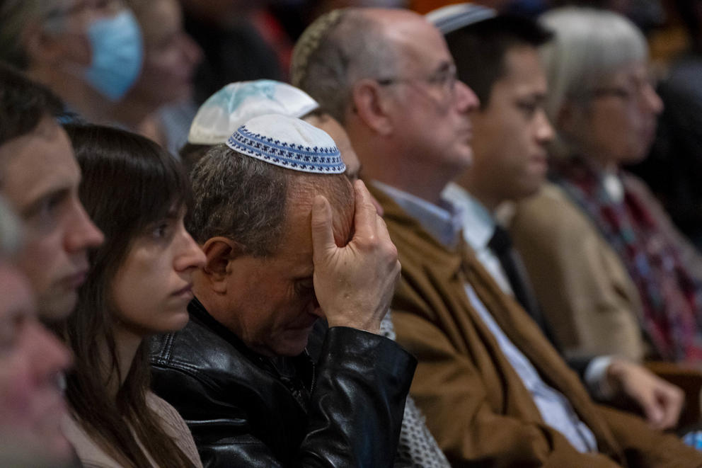 A man wearing a yarmulke covers his face with his hand as he sits in a crowded pew in a synagogue