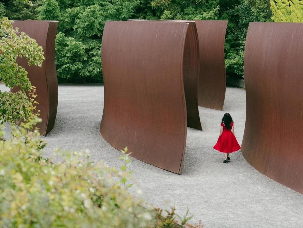 photo of a large sculptural outdoor installation that looks like rust colored waves, a woman in a red dress walks thru