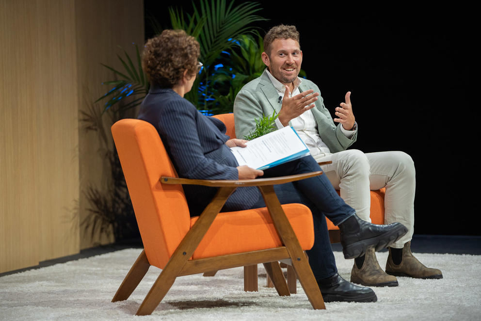 a man and woman sit in orange chairs on a stage having a conversation