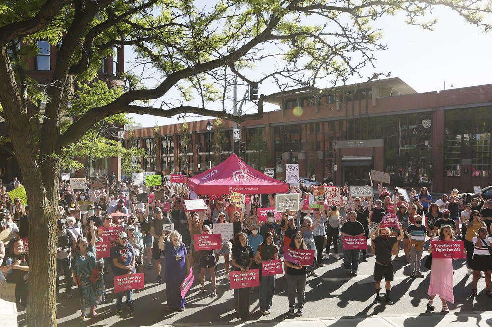 People holding signs with slogans supporting a woman's right to choose an abortion stand in front of a building and a Planned Parenthood tent.