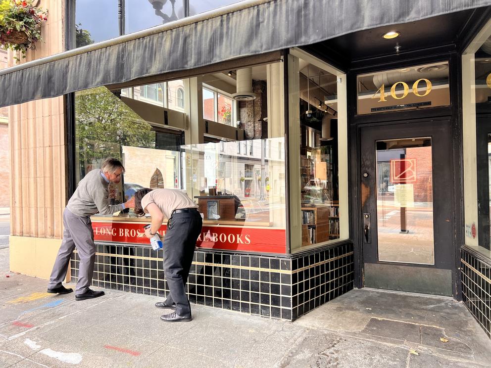 two people on a sidewalk clean the storefront window of a bookstore