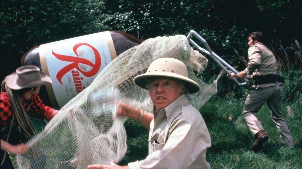 film still of an older man in a classic safari suit attempting to capture a giant Rainier Beer in a net